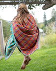 A young barefoot girl with loose blonde hair sits on a swing over green grass wrapped in a flannel blue and red camp blanket. 